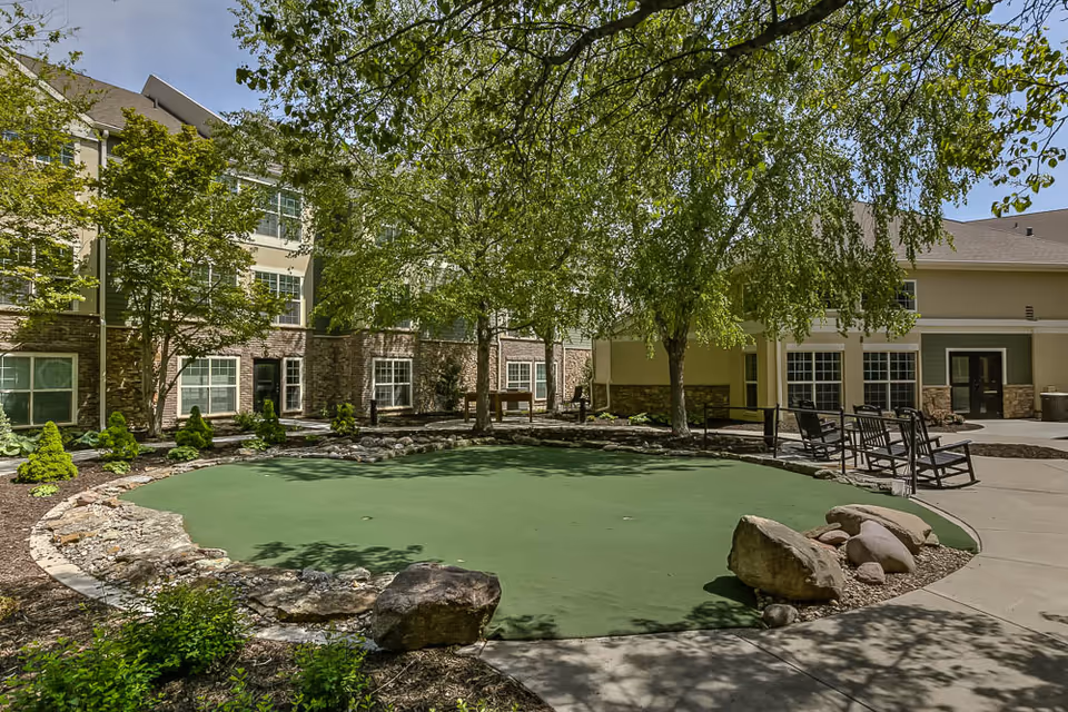 Outdoor courtyard area at Remington Heights Retirement Community featuring a putting green surrounded by rocks and landscaping. There are several trees providing shade, benches for seating, and a multi-story building with large windows in the background.