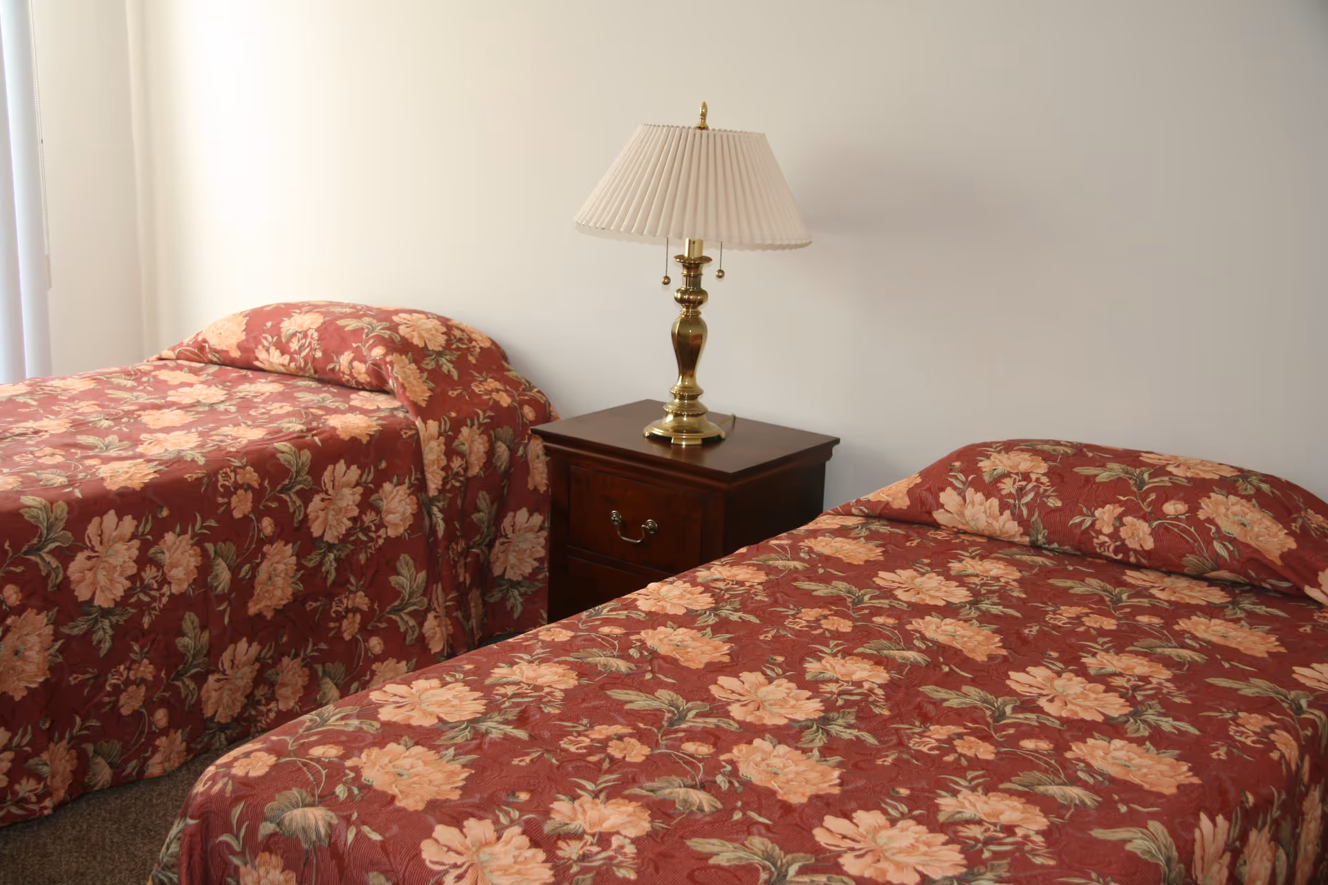 Two twin beds with matching red floral bedspreads and a nightstand with a brass lamp in a simple bedroom.