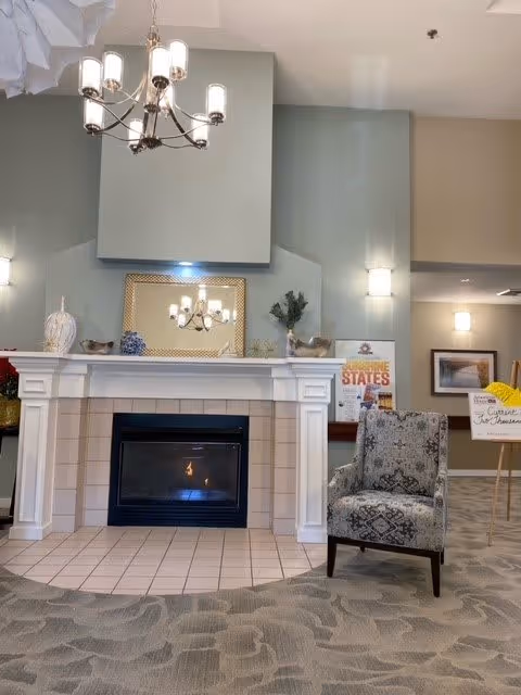 A cozy interior space featuring a lit fireplace with a white mantel, a decorative mirror above it, and a patterned armchair to the right. The walls are painted light gray, and there is a chandelier hanging from the ceiling. Various decorative items and framed pictures are placed around the fireplace area.