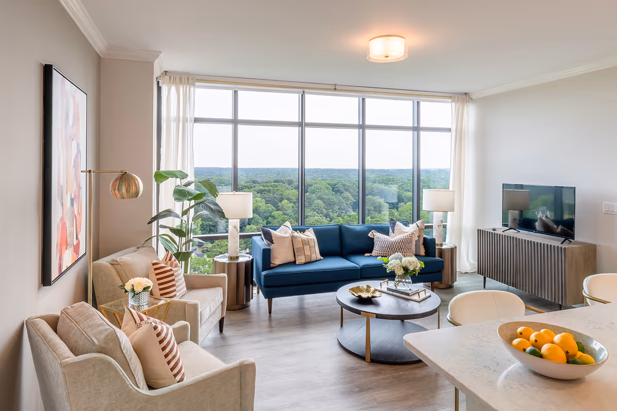 A bright and modern living room with large floor-to-ceiling windows offering a scenic view of a green forest. The room features a blue sofa with decorative pillows, two beige armchairs with striped pillows, a round coffee table with a flower arrangement, two side tables with lamps, a flat-screen TV on a wooden console, and a kitchen counter with a bowl of fruit in the foreground.