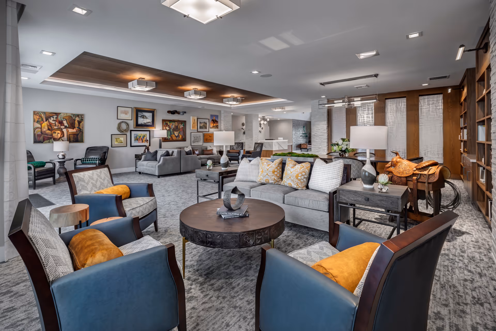 A spacious and well-lit living room area in a senior living facility featuring comfortable seating including a gray sofa with patterned pillows, blue armchairs with orange cushions, a round wooden coffee table, side tables with lamps, and various framed artworks on the walls. The room has a modern ceiling design with recessed lighting, a bookshelf, and a decorative saddle on display.