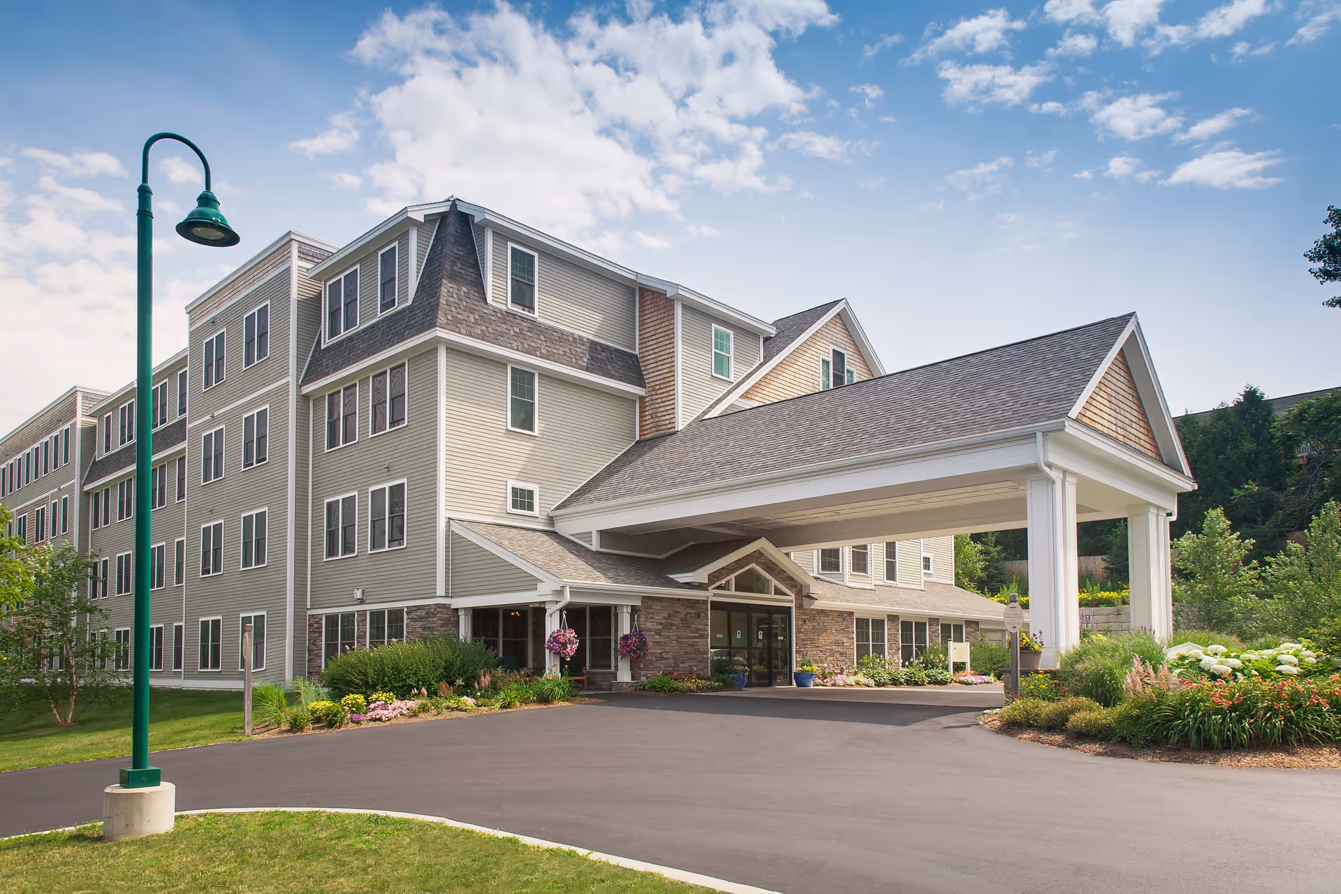 Exterior view of a multi-story senior living facility building with beige siding and stone accents. The building has many windows and a large covered entrance with white pillars. There is a paved driveway and landscaped greenery with flowers around the entrance. A green street lamp is visible on the left side under a partly cloudy sky.