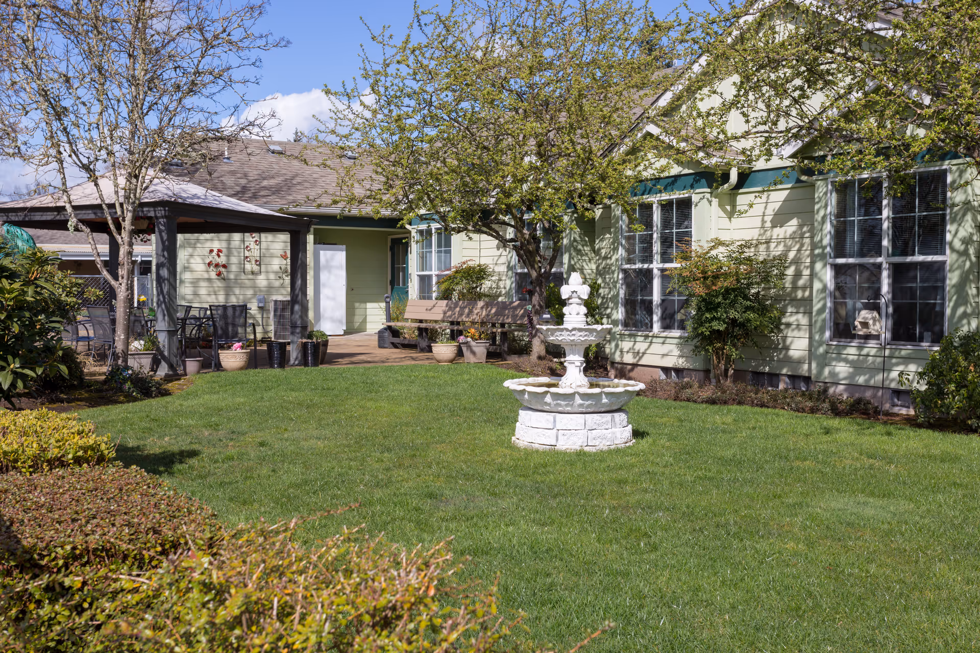 Courtyard featuring a white tiered fountain on a green lawn, trees, potted plants, and a light-green building with large windows and a covered patio.