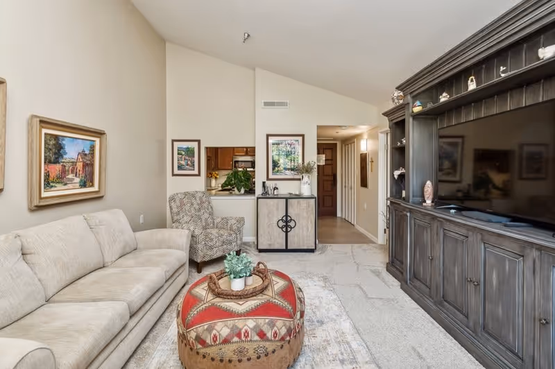 A cozy living room with a beige sofa, patterned armchair, and a large wooden entertainment center with a flat-screen TV. A round ottoman with a red and beige patterned cover sits in the center, topped with a small plant and decorative tray. The walls are adorned with framed artwork, and a small cabinet with decorative items is placed against the far wall near an open doorway leading to a kitchen area.
