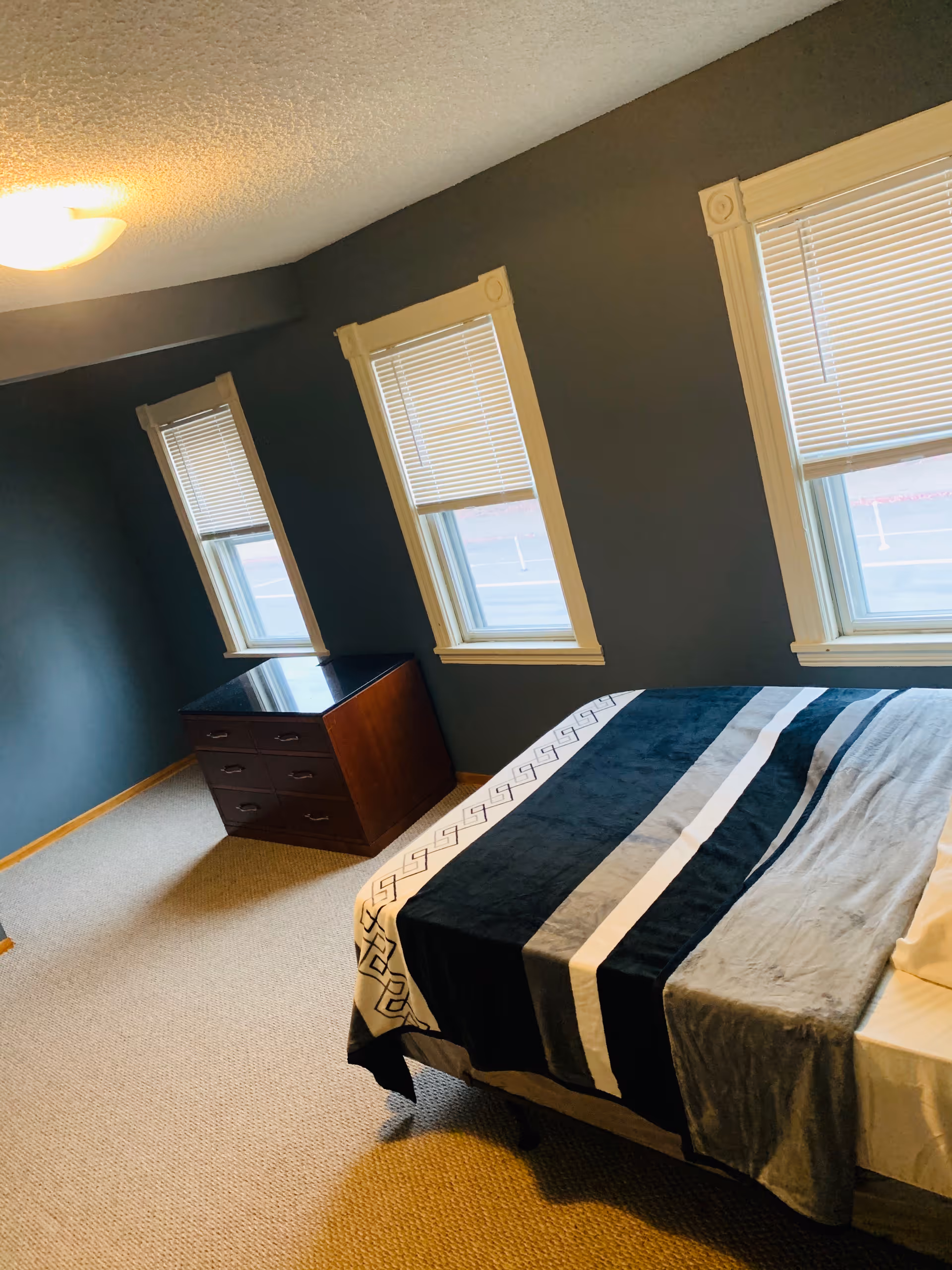 A bedroom with dark gray walls and beige carpet. There are three windows with white blinds partially closed. A bed with a striped blanket in shades of black, gray, and white is positioned near the windows. A wooden dresser with a dark top is placed against the wall under one of the windows. The room is lit by a ceiling light.