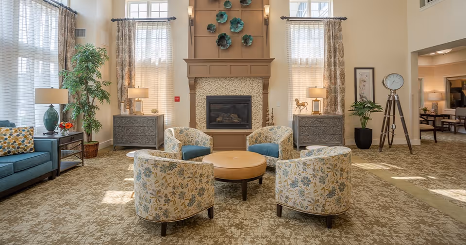 Bright communal living room with a central fireplace, four patterned armchairs around a round ottoman, sofa, tall windows, and decorative accents.