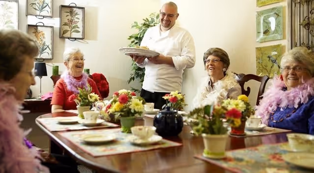 A group of elderly women sitting around a dining table decorated with flowers and tea cups, smiling and wearing colorful feather boas, while a man in a white chef's coat serves a plate of food.