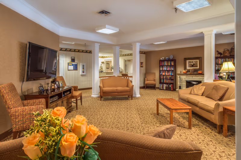 A cozy living room area in a senior living facility with beige walls and patterned carpet. The room features multiple seating options including sofas and armchairs, a wooden coffee table, a flat-screen TV mounted on the wall, bookshelves filled with books, a fireplace decorated with pumpkins, and a lamp on a side table. There are white columns dividing the space and a bouquet of orange roses in the foreground.