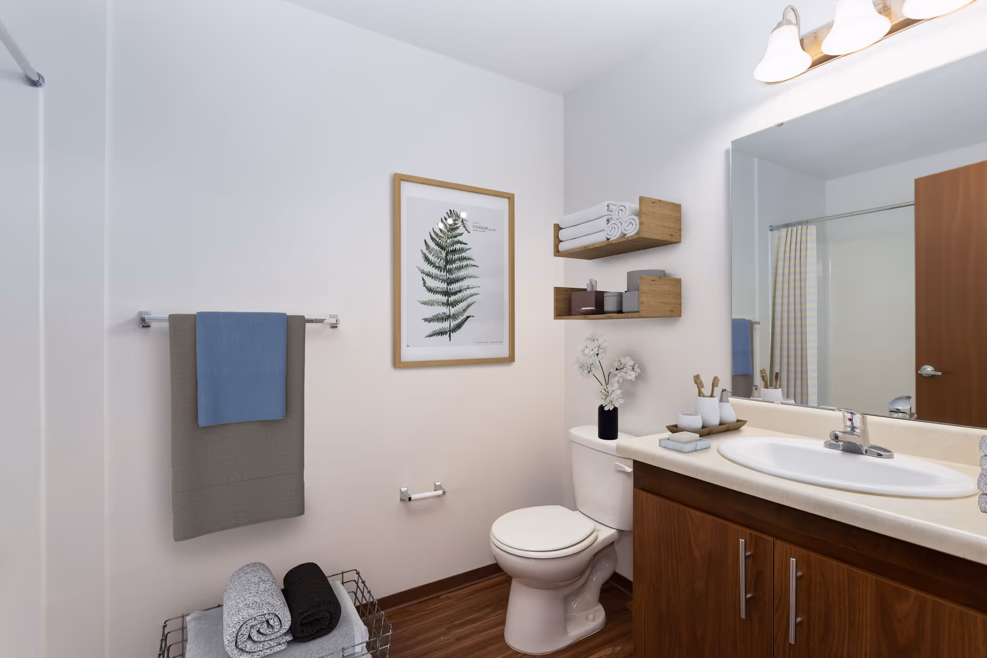 A clean and modern bathroom with a white toilet, wooden vanity with a white sink, and a large mirror above. There are two wooden shelves mounted on the wall holding rolled white towels and small containers. A framed botanical print hangs on the wall above a towel rack with a gray and blue towel. A wire basket on the floor holds rolled towels. The bathroom has wood flooring and a closed wooden door is visible in the mirror reflection.