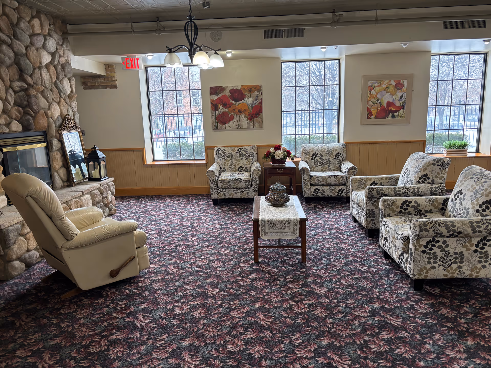 A cozy assisted-living lounge with patterned armchairs and a recliner arranged around a coffee table near a stone fireplace and large windows.