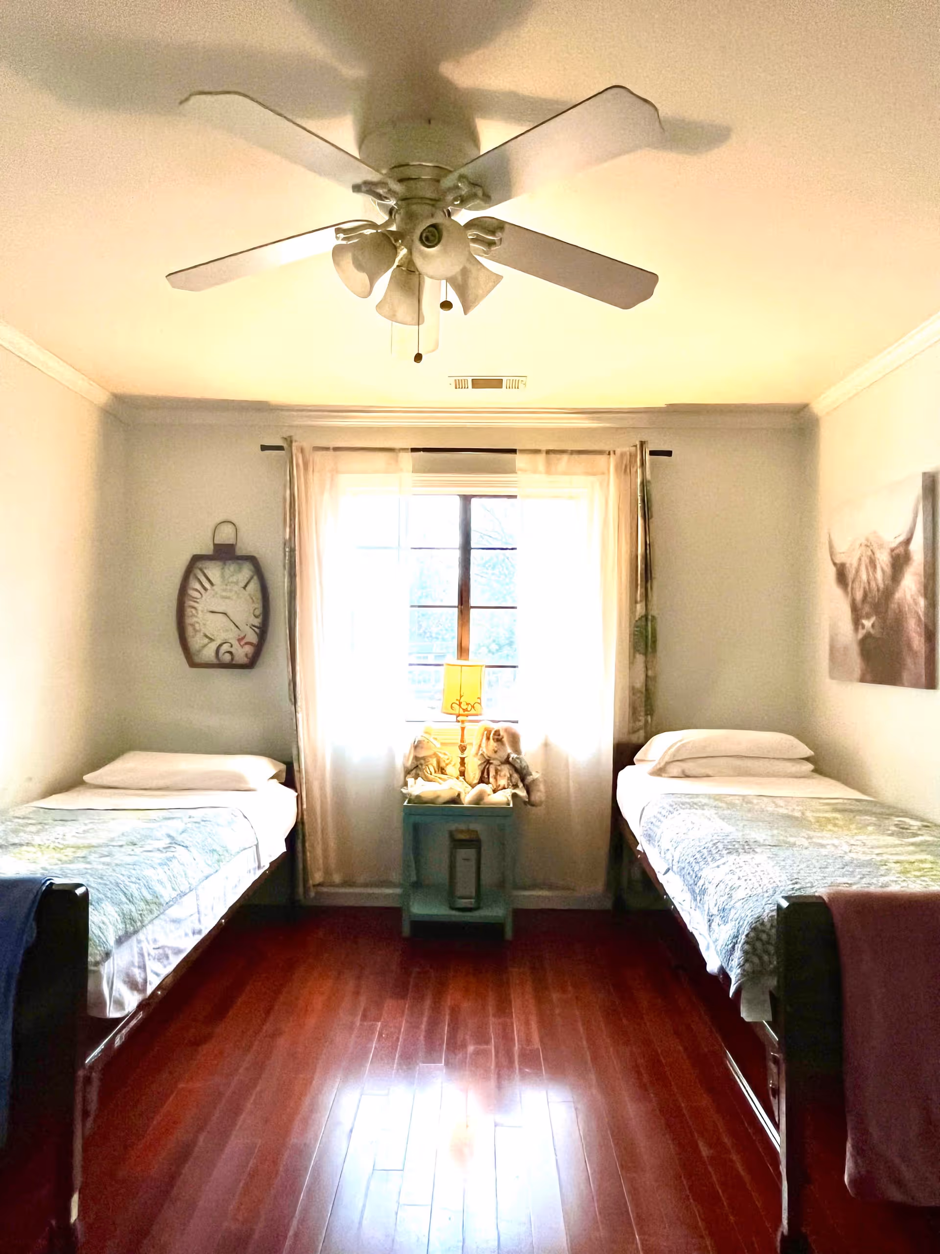 Bright shared bedroom with two single beds separated by a small nightstand and window, hardwood floor, and a ceiling fan.