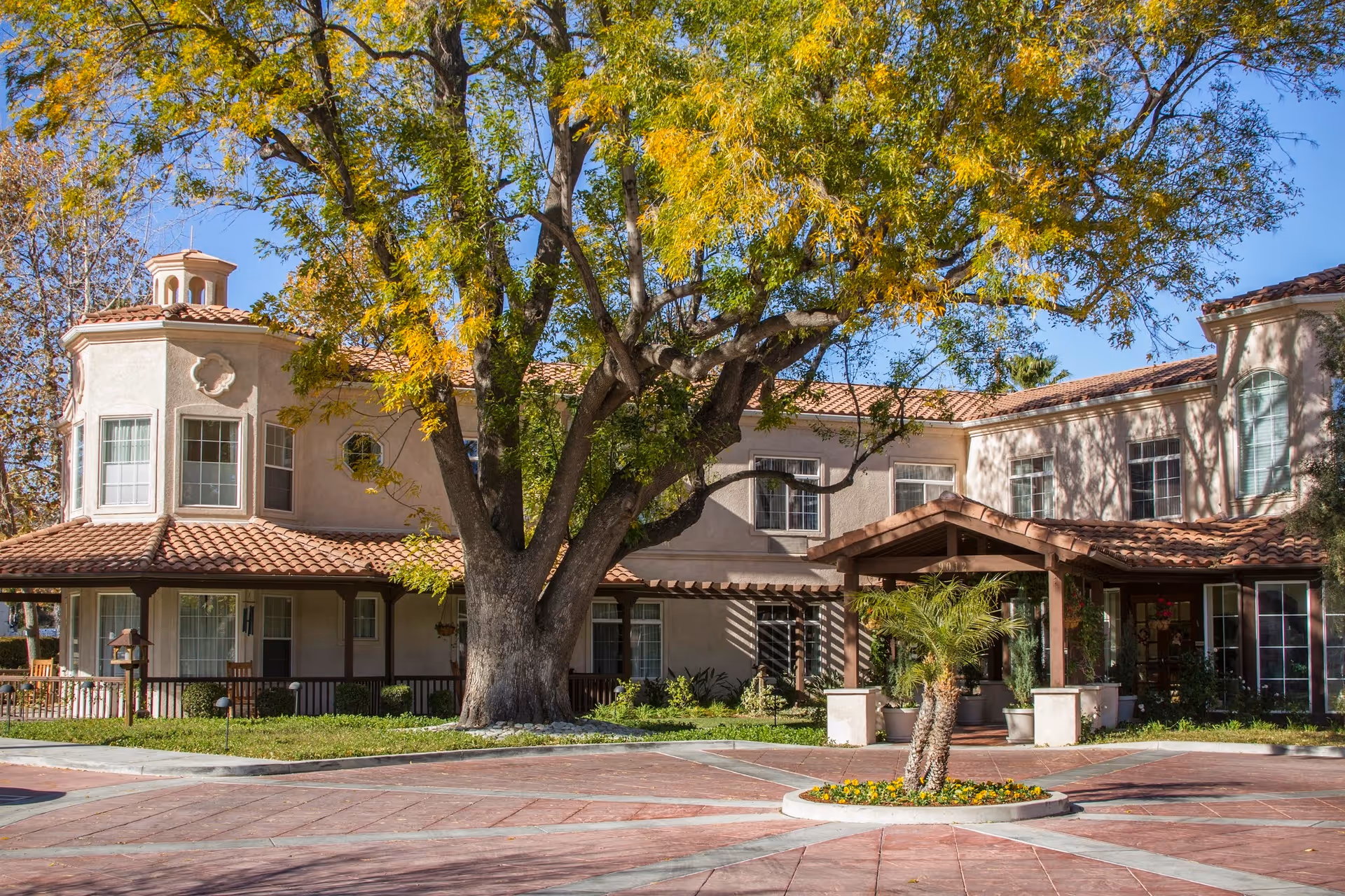 Exterior view of a two-story senior living facility building with beige stucco walls and a red tile roof. A large tree with yellow and green leaves stands prominently in front of the building near the entrance, which has a covered porch supported by wooden beams. There is a circular driveway with a small landscaped island featuring a palm tree and flowers.