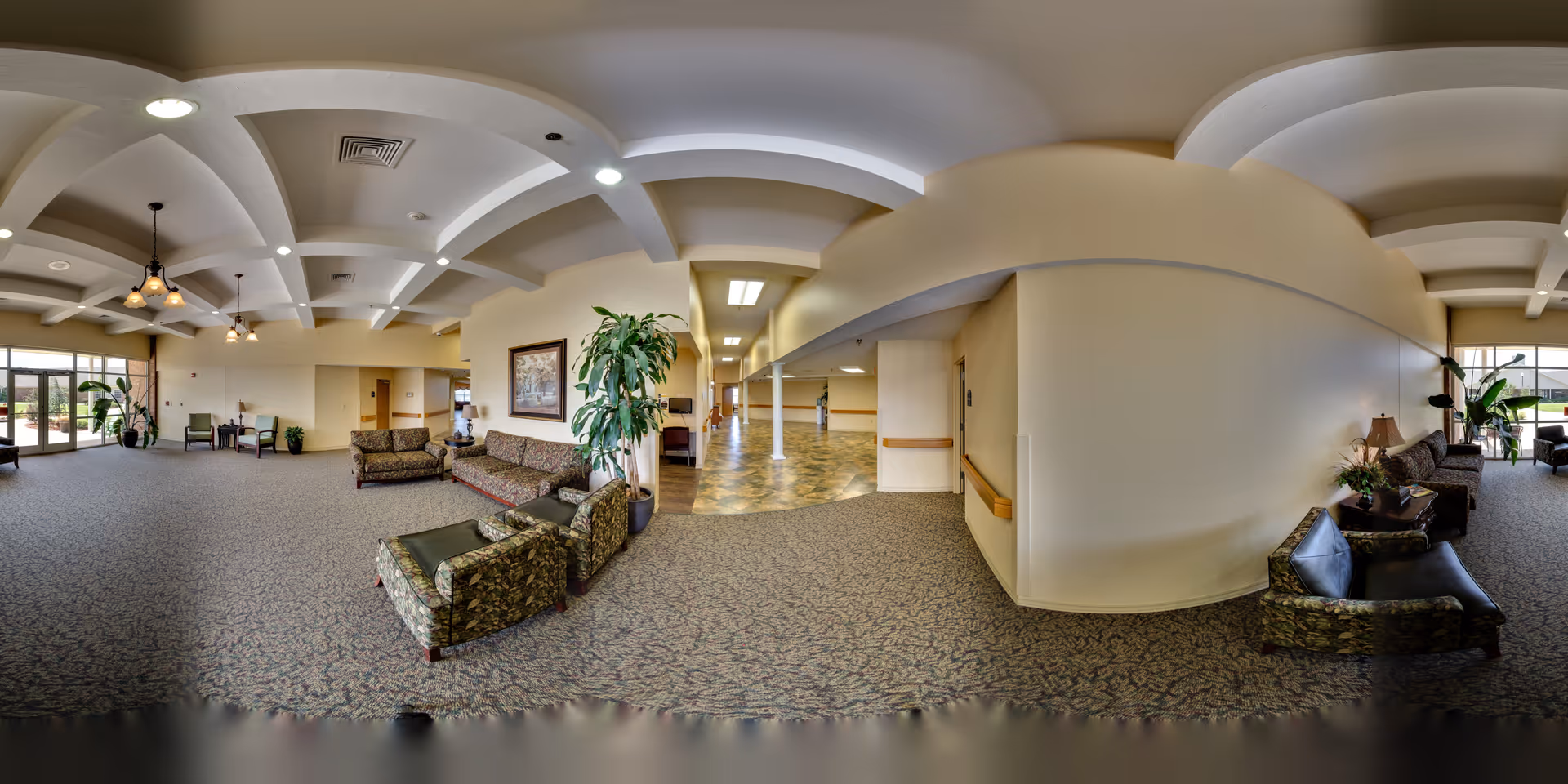 A spacious and well-lit common area in a senior living facility with patterned carpet flooring, multiple seating arrangements including sofas and armchairs, potted plants, framed artwork on the walls, and large windows allowing natural light to enter. The ceiling features decorative beams and hanging light fixtures. A hallway with tiled flooring extends from the center of the room.