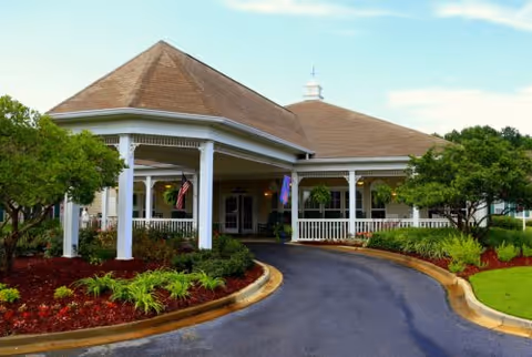 Exterior view of a senior living facility with a large covered entrance, white columns, and a brown roof. The driveway curves in front of the entrance, surrounded by well-maintained landscaping with green shrubs, trees, and red mulch. The sky is clear with some clouds.