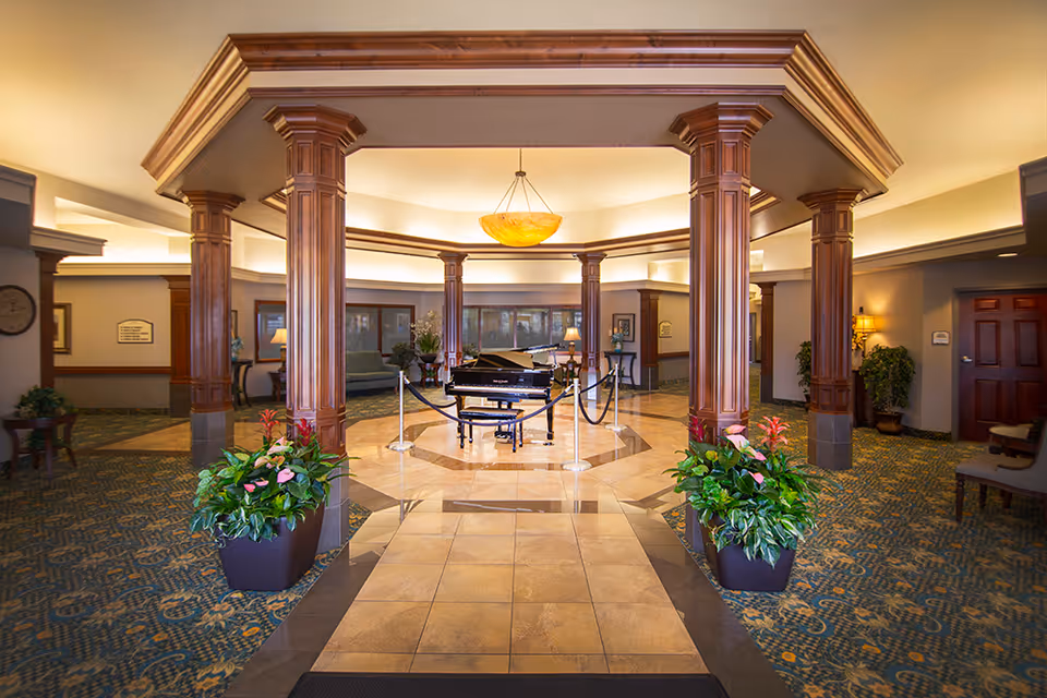 Spacious and elegant interior lobby area with a grand piano in the center surrounded by wooden columns and roped stanchions. The floor features a combination of tiled and carpeted areas, with large potted plants on either side of the tiled walkway. Comfortable seating and lamps are visible in the background, along with warm lighting from a ceiling fixture.