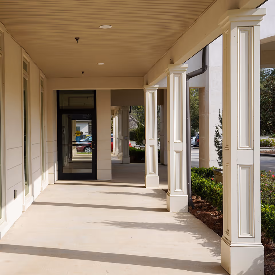 Covered walkway outside a building with cream-colored columns and a beige ceiling, leading to a glass door entrance. There are bushes and parked cars visible in the background.