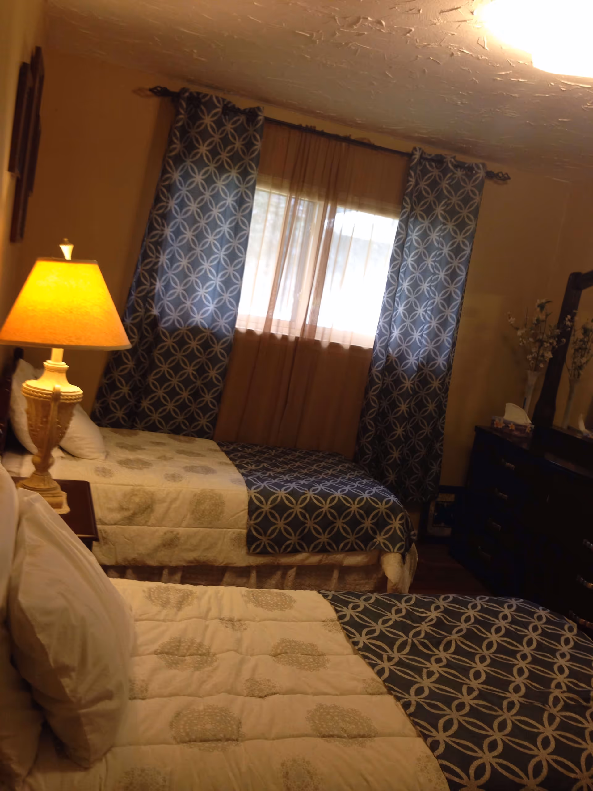 A bedroom with two twin beds covered in matching beige and blue patterned bedding. A wooden nightstand with a beige lamp is positioned between the beds. A window with sheer and patterned blue curtains is centered on the far wall. To the right, there is a dark wooden dresser with a mirror and decorative items on top.