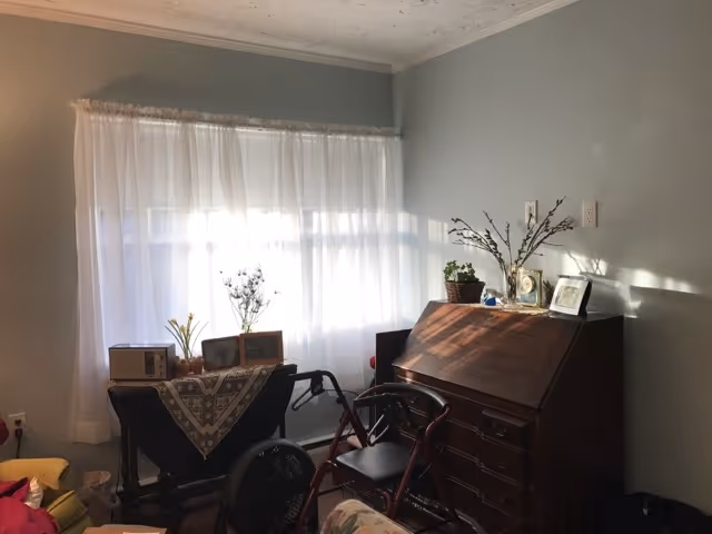 A cozy room with light gray walls and a window covered by sheer white curtains letting in sunlight. There is a wooden desk with drawers and decorative items on top, including a plant and framed pictures. In front of the desk is a red walker with a black seat. To the left, there is a small table covered with a patterned cloth, holding a microwave, a fan, and some other small items. Part of a yellow armchair and a floral-patterned sofa are also visible.
