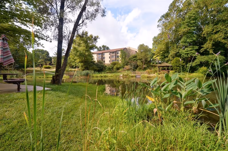 Grassy landscaped grounds with a pond, trees, and a multi-story building in the background under a partly cloudy sky.