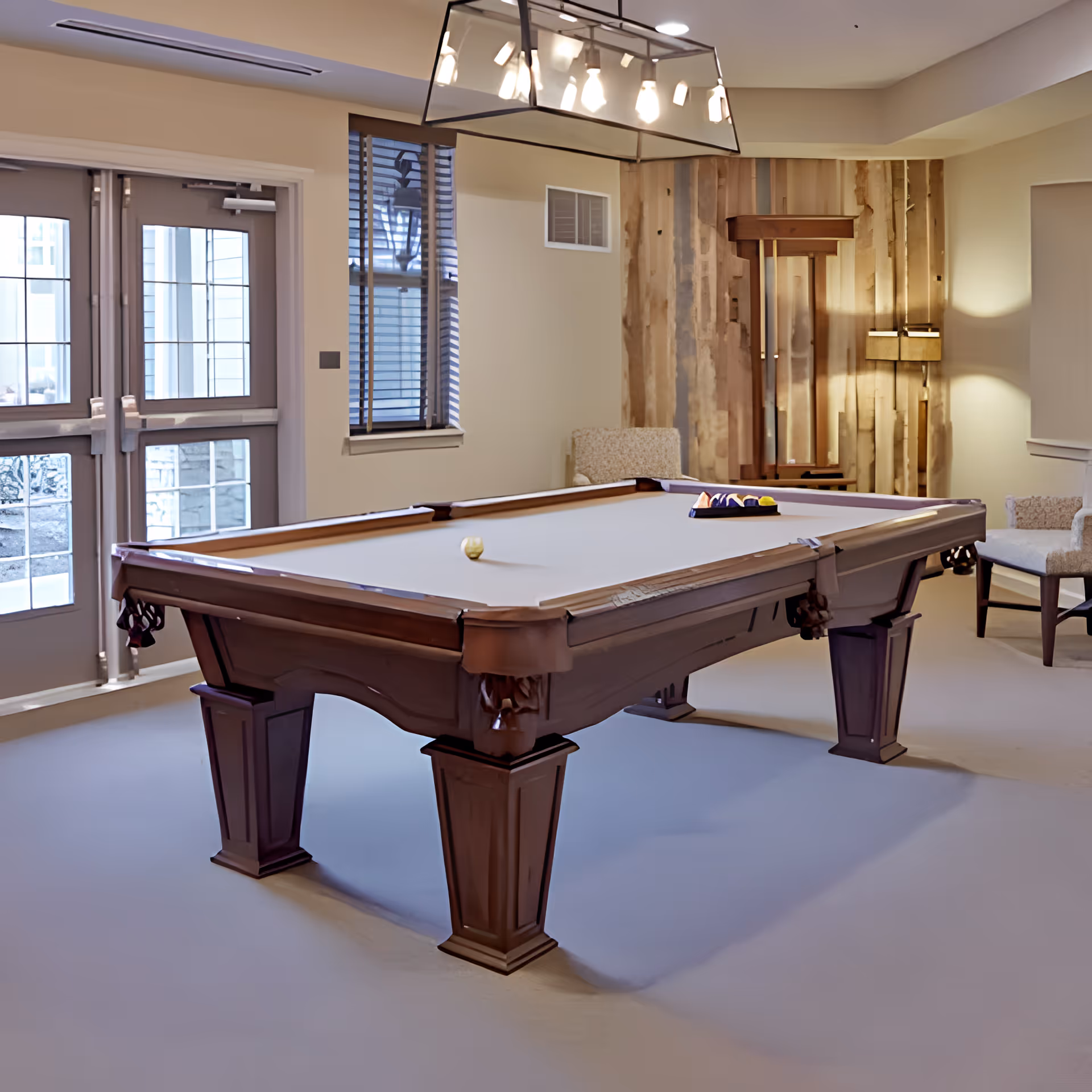 A well-lit indoor recreational room featuring a wooden pool table with billiard balls arranged on it. The room has beige walls, a window with blinds, two upholstered chairs, and a modern light fixture hanging above the pool table. There is a wooden decorative panel on one wall and double glass doors leading outside.