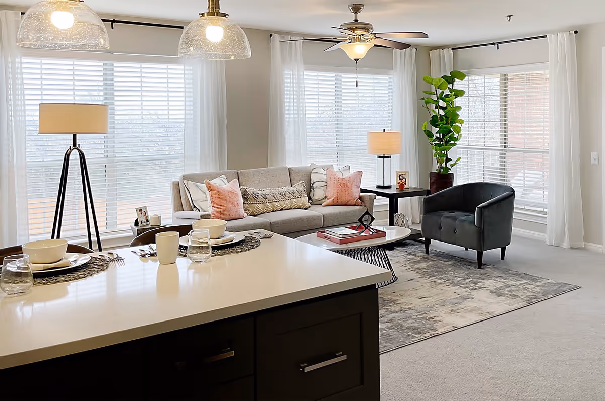 Bright and modern living room with large windows covered by white blinds and sheer curtains. The room features a gray sofa with pink and patterned pillows, a black armchair, a coffee table with books and decor, a floor lamp, a side table with a lamp and photo frame, a tall green plant, and a kitchen island with place settings including bowls, plates, cups, and glasses.