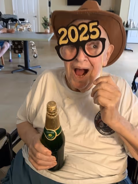 An elderly person wearing a brown cowboy hat and holding novelty glasses with '2025' on them, smiling and holding a bottle with a gold foil top, seated in a wheelchair inside a room with tables and chairs.