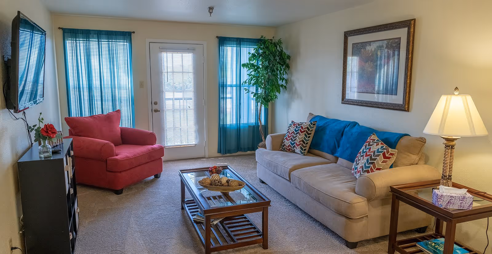A cozy living room with a beige sofa adorned with colorful patterned pillows and a blue throw blanket. Next to the sofa is a wooden side table with a lamp and a tissue box. Across from the sofa is a red armchair beside a black shelving unit with a vase holding a red rose. The room has a glass coffee table with decorative items on it, beige carpet, and two windows with teal curtains flanking a glass door with blinds. A framed picture hangs on the wall above the sofa, and a tall green plant is placed near the window.