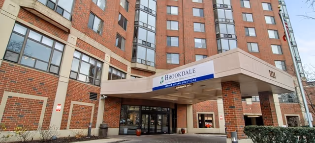 Exterior view of the entrance to Brookdale Oak Park senior living facility, showing a multi-story brick building with large windows and a covered drop-off area with a sign that reads 'Brookdale Senior Living Solutions'.