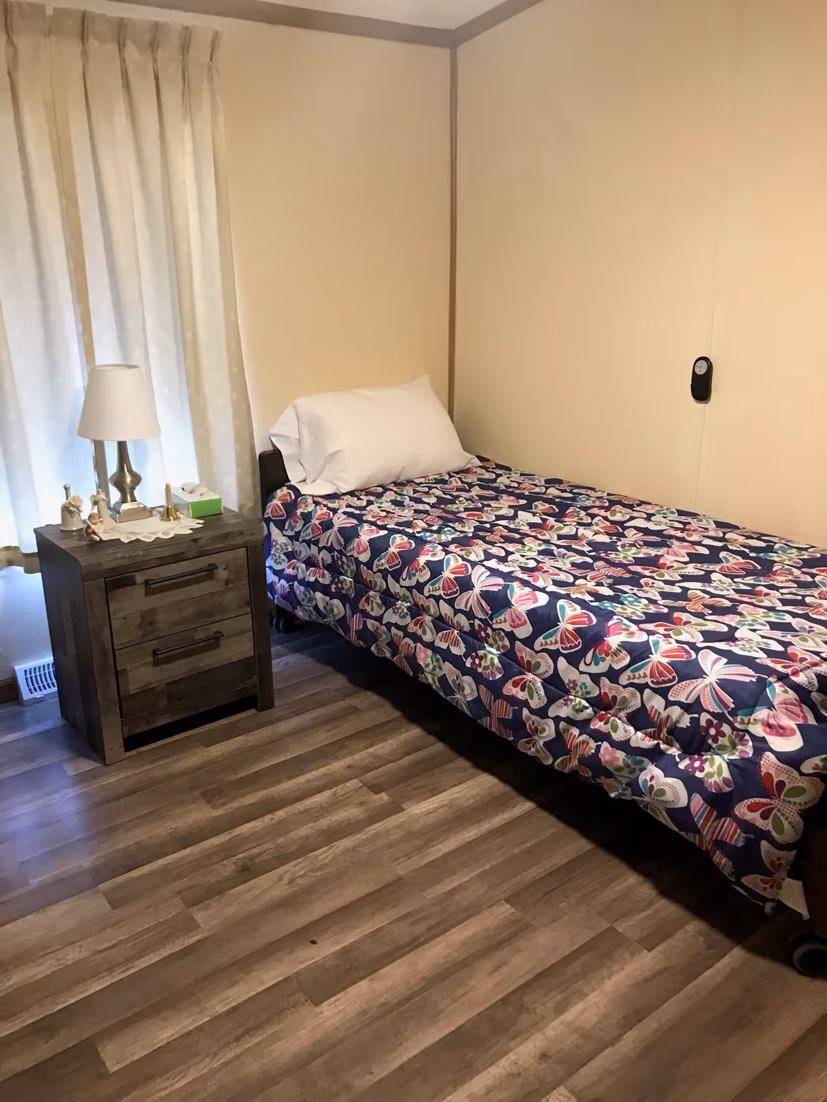 A small bedroom with a single bed covered with a colorful butterfly-patterned blanket and a white pillow. Next to the bed is a wooden nightstand with two drawers, a white table lamp, a tissue box, and small decorative figurines. Beige curtains cover the window, and the floor is wood laminate.