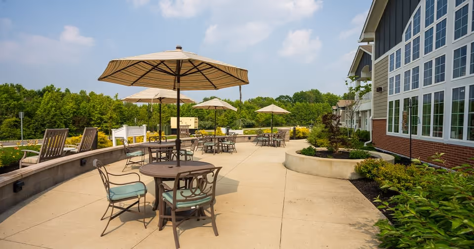 Outdoor patio area at Juniper Village at Hamilton with multiple round tables and chairs under large beige umbrellas, surrounded by greenery and a building with large windows on the right side.