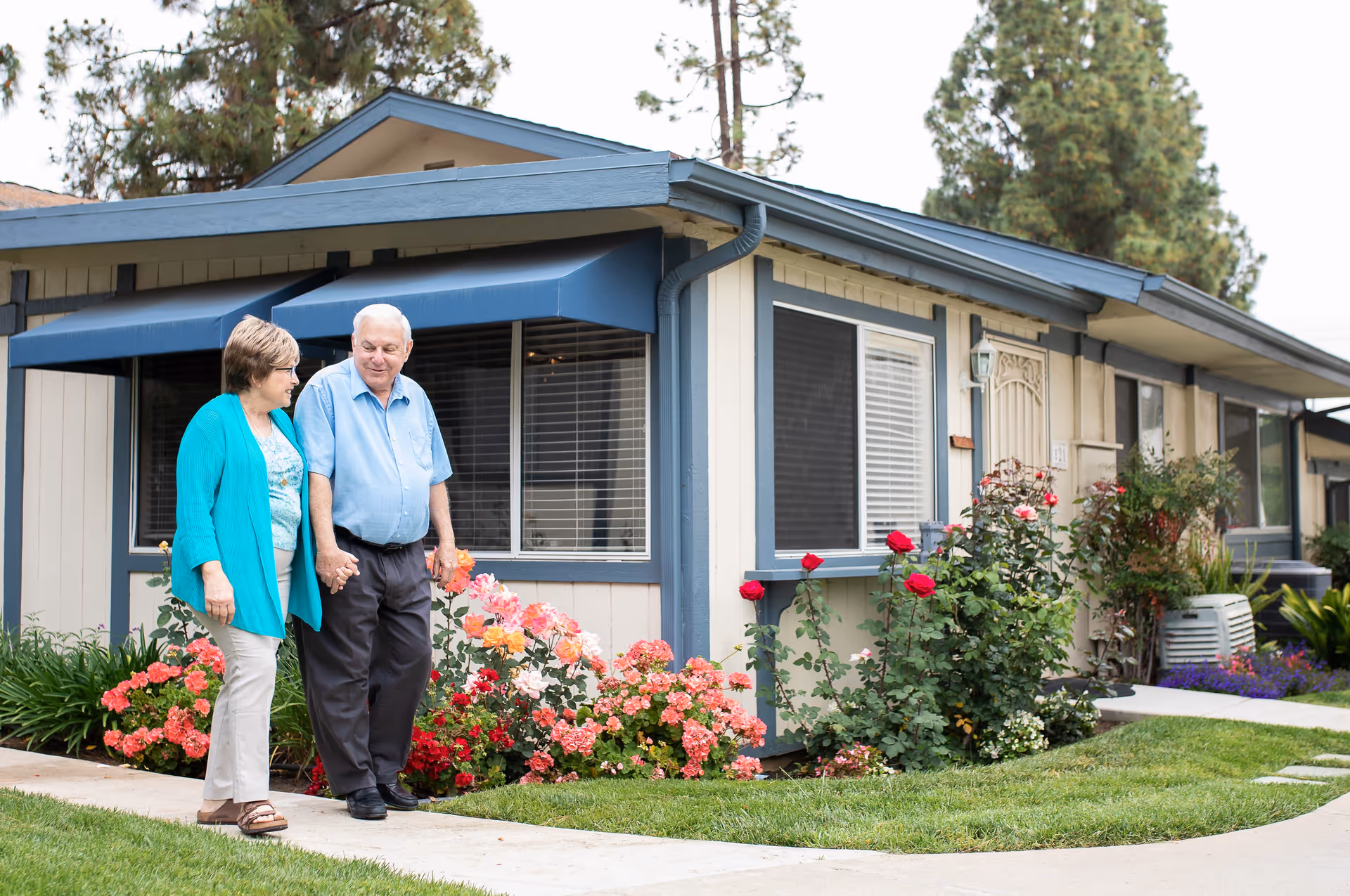 An elderly couple holding hands and walking on a sidewalk in front of a single-story residential building with blue trim and an awning. There are colorful flowers and green grass along the walkway.