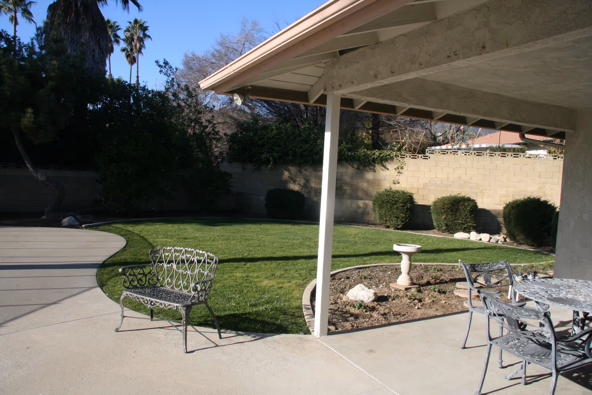Outdoor patio area with metal chairs and a table under a covered porch, a small garden bed with a birdbath, green lawn, bushes, and a brick wall surrounding the yard with trees in the background.