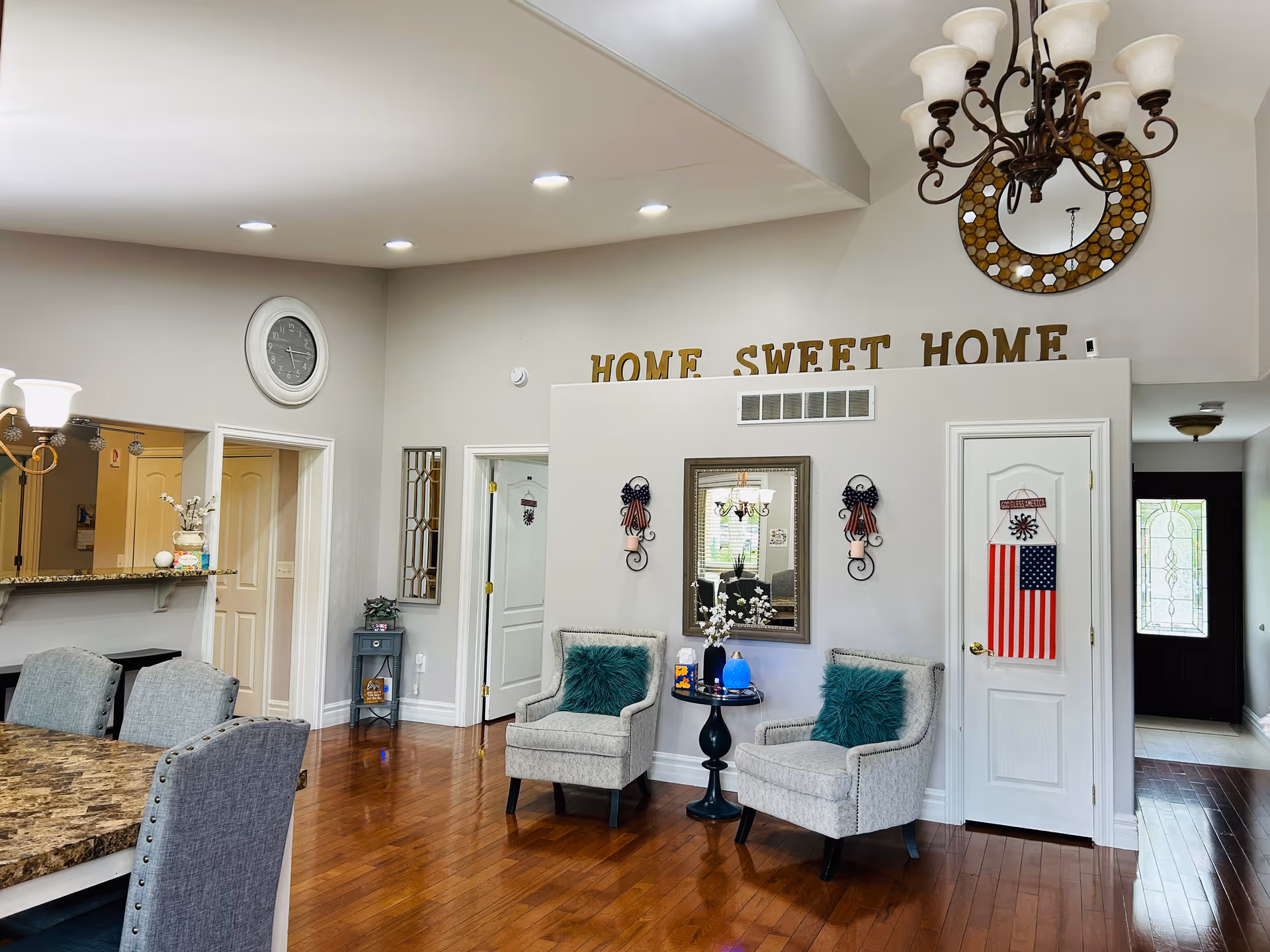 A cozy interior living area with two gray upholstered chairs with teal pillows, a small round black table with decorative items, a wall mirror, and wall sconces. Above the wall is a decorative sign that reads 'HOME SWEET HOME'. To the left is a dining area with a granite countertop and gray upholstered chairs. The floor is wooden, and the walls are painted light gray. A chandelier hangs from the ceiling, and an American flag decoration is on a white door to the right.