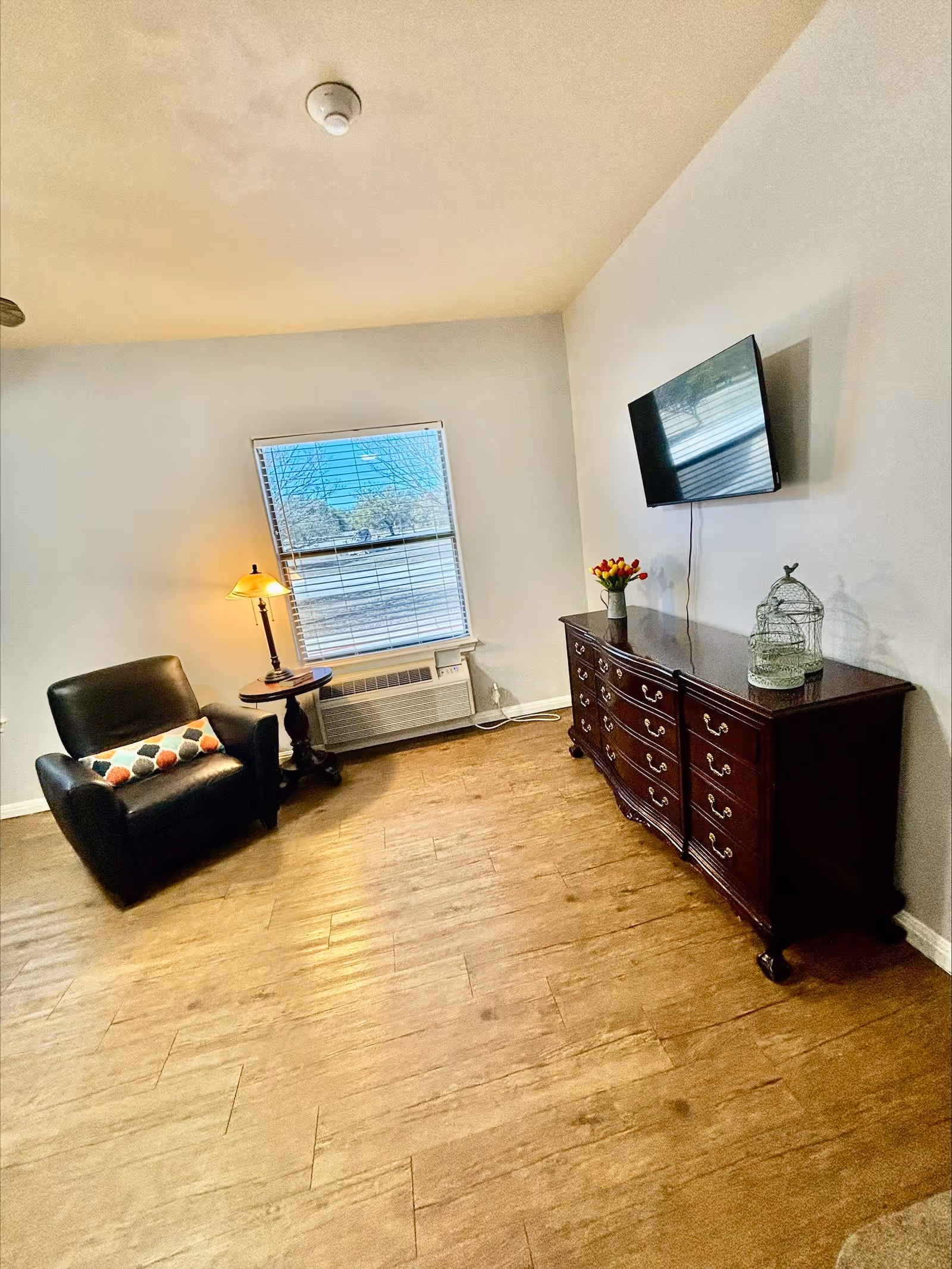 A cozy living room area with a black leather armchair featuring a colorful pillow, a small round wooden side table with a lamp, a window with blinds, a wall-mounted flat-screen TV, and a dark wooden dresser decorated with a vase of flowers and a decorative birdcage.