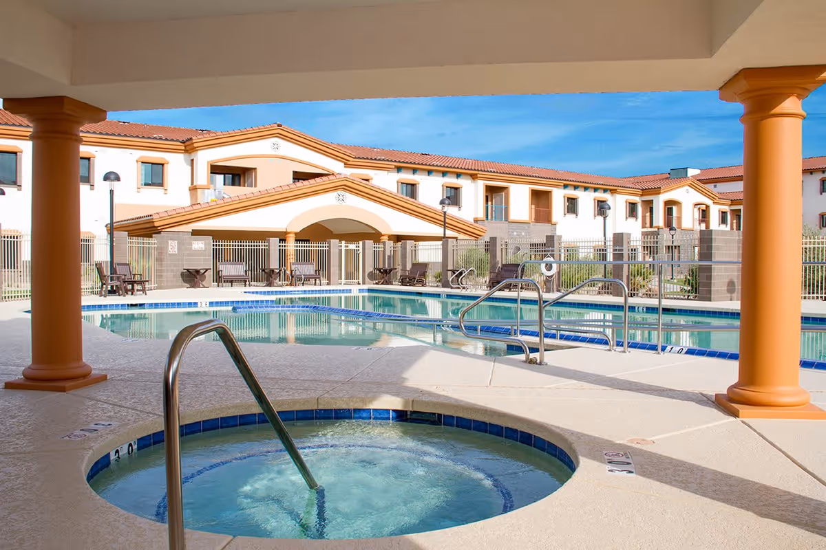 Outdoor swimming pool and hot tub area at Fellowship Square Surprise, with a covered patio supported by columns in the foreground and a two-story building with a tiled roof in the background under a clear blue sky.
