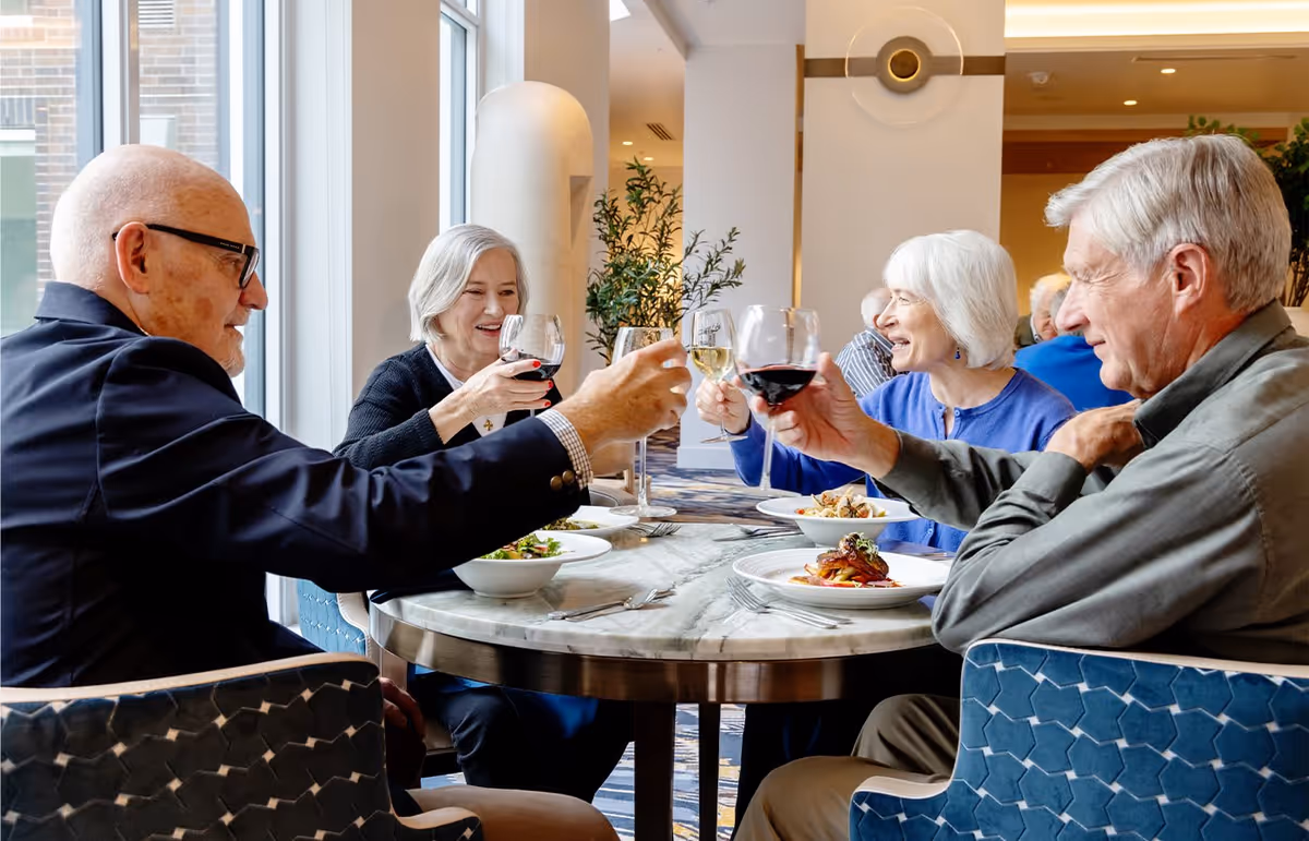 Four elderly people sitting around a round table in a dining area, raising their wine glasses in a toast. Plates of food are on the table, and large windows let in natural light. The setting appears warm and social.