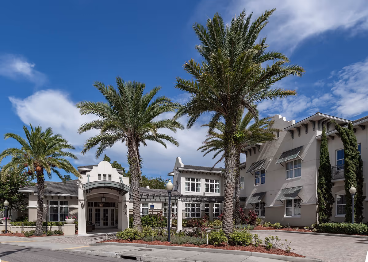 Front exterior of a senior living building with palm trees, an arched entrance, and a clear blue sky.