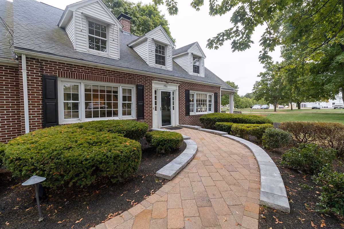 Brick residential-style building entrance with a curved paved walkway, manicured shrubs, and dormer windows.