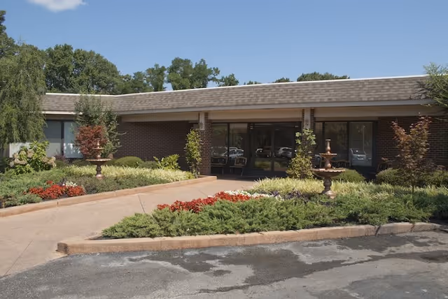 Exterior view of a single-story brick building with a sloped roof, surrounded by landscaped greenery and flower beds. There is a paved walkway leading to the glass entrance doors, with two decorative fountains on either side of the walkway. Trees and shrubs are visible around the building under a clear blue sky.