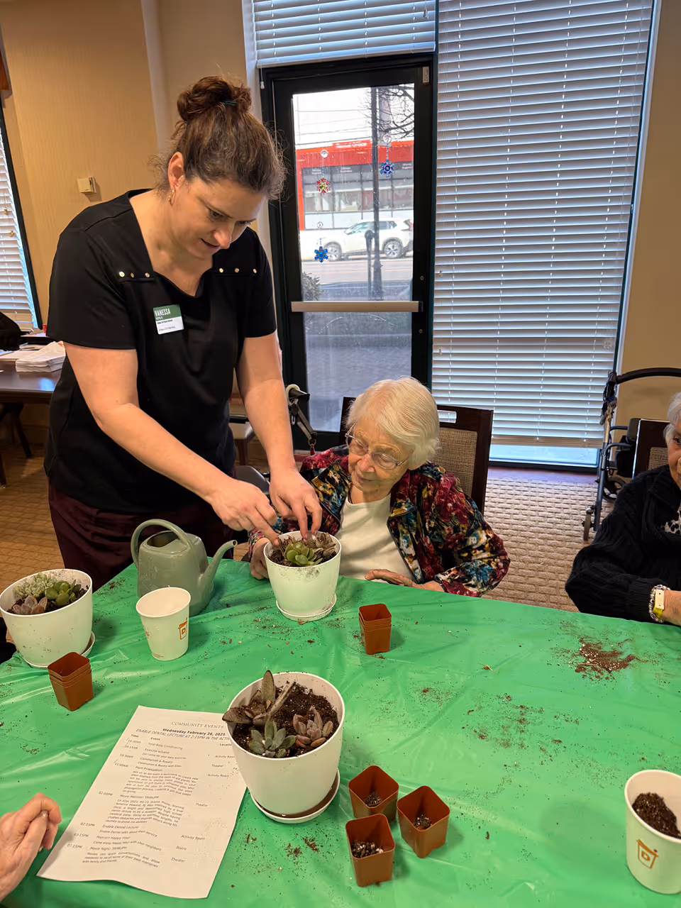 A woman assisting an elderly woman with planting succulents in white pots at a table covered with a green plastic tablecloth. There are small brown pots, a watering can, and a paper with community events on the table. The setting appears to be indoors with large windows and blinds in the background.