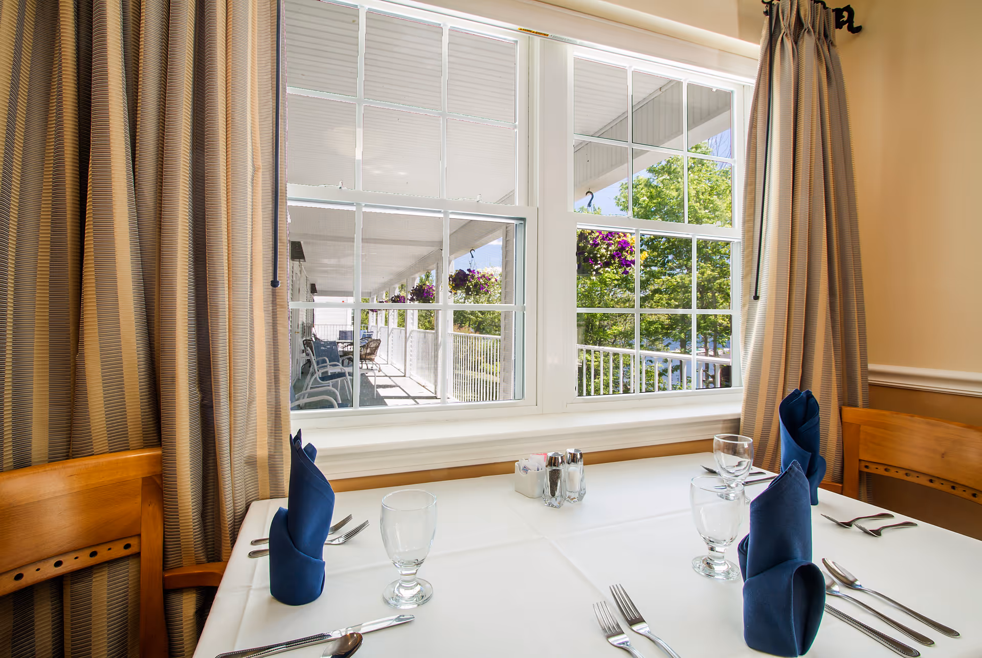 A dining table set for four with white tablecloth, blue folded napkins, glasses, and silverware near a window with beige curtains. Outside the window, a covered porch with hanging flower baskets and outdoor seating is visible.