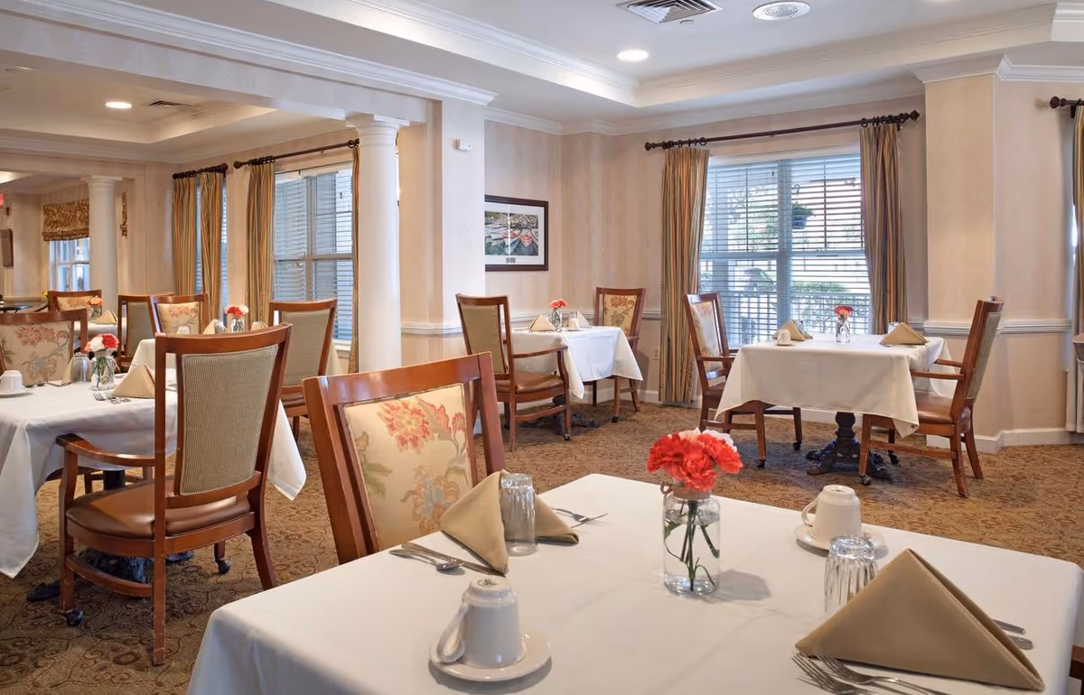 Dining room with several tables set with white tablecloths, napkins, floral centerpieces, and wooden chairs.