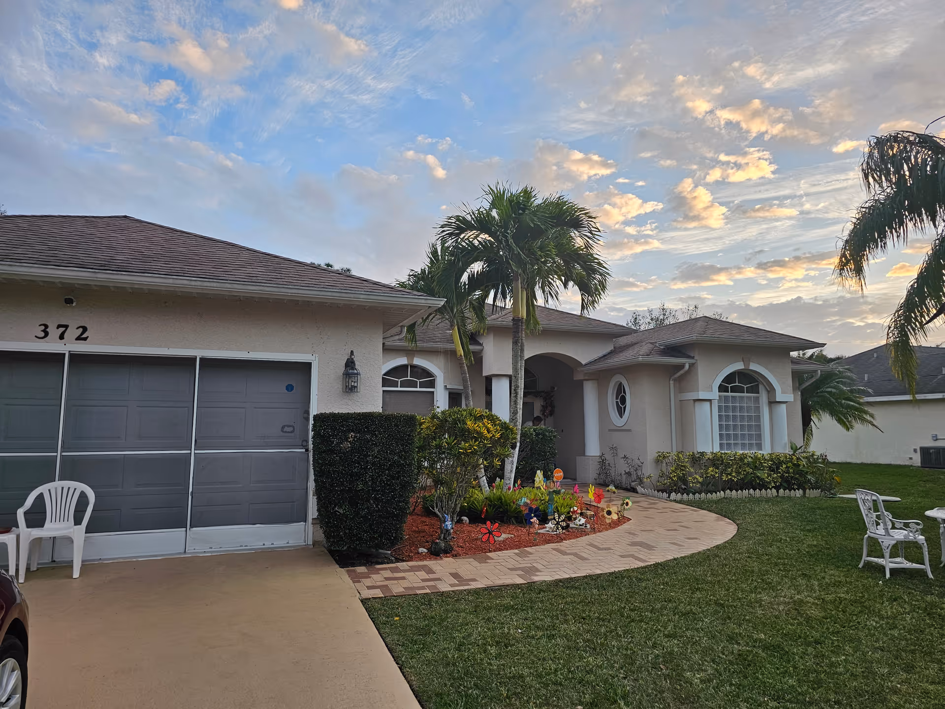 Exterior view of a single-story residential building with a garage, a paved walkway, palm trees, and a well-maintained lawn under a partly cloudy sky during sunset.