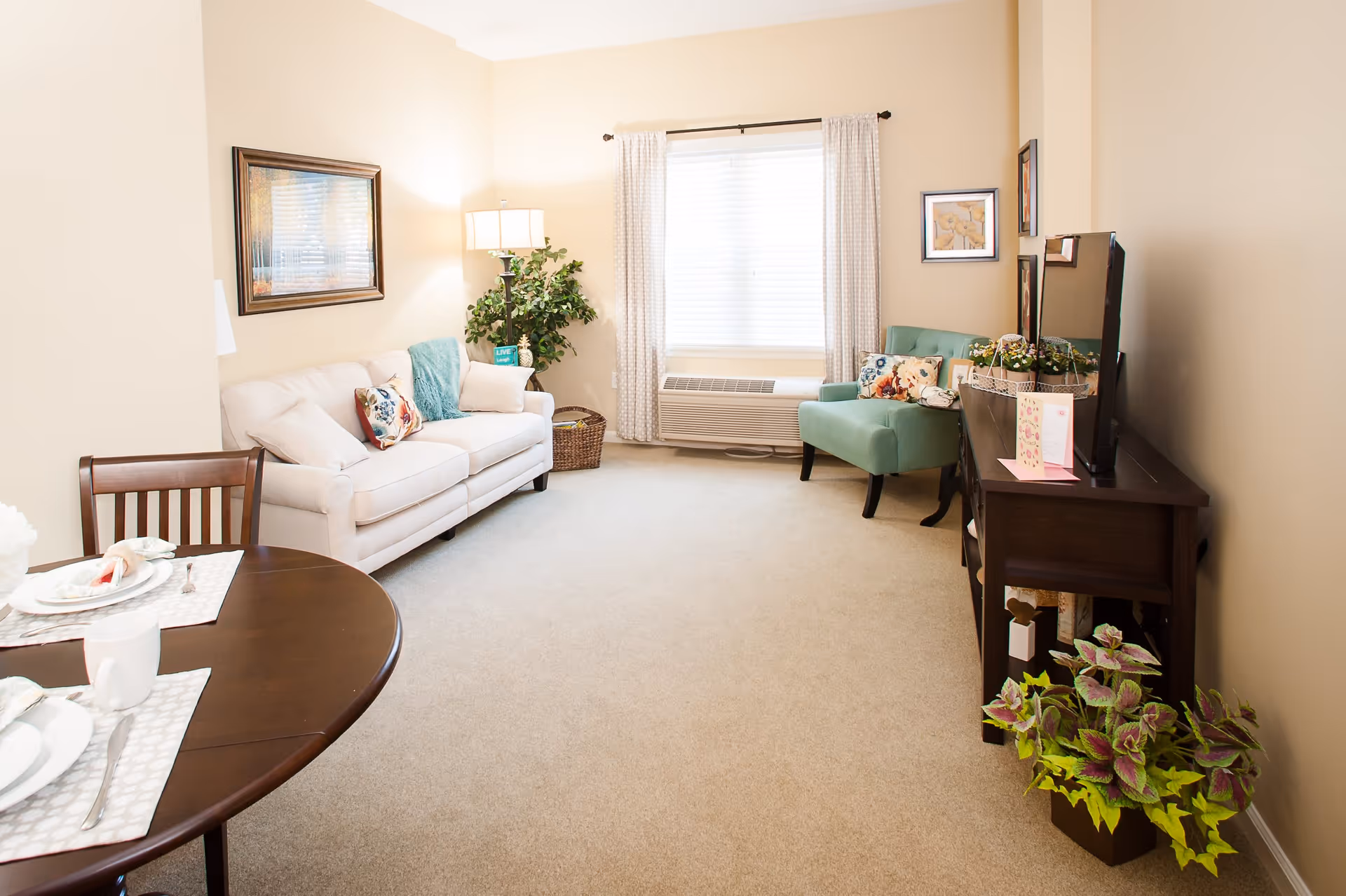A bright and cozy living room with beige walls and carpet. The room features a white sofa with decorative pillows and a teal throw blanket, a green armchair with floral pillows, a dark wooden TV stand with a flat-screen TV, and a round wooden dining table set with white plates and cups. There is a floor lamp and a potted plant near the window with white curtains and blinds.