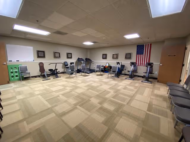 A spacious room with exercise equipment arranged along the walls, including stationary bikes and weight machines. The room has beige patterned carpet, a whiteboard on one wall, framed pictures, and an American flag hanging on the far wall. Several chairs are lined up against the walls.