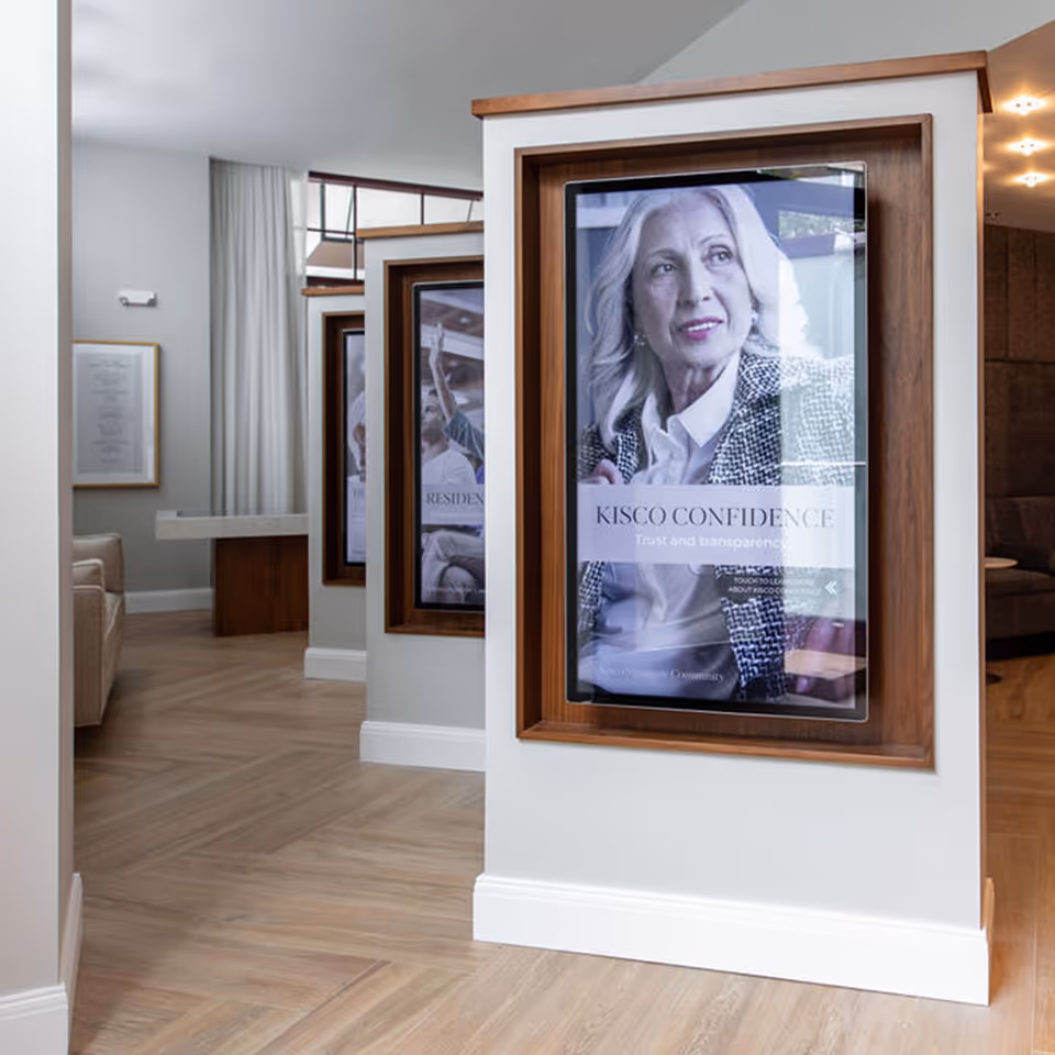 Interior view of a senior living facility hallway with wooden framed digital displays mounted on white walls. The closest display shows a portrait of an older woman with the text 'KISCO CONFIDENCE Trust and transparency.' The hallway has light wood flooring and neutral-colored walls with a glimpse of seating areas and curtains in the background.