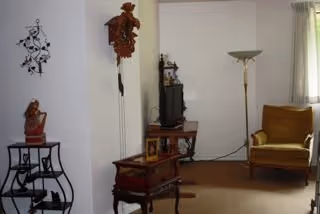 A cozy living room with a mustard yellow armchair near a window with light curtains, a tall floor lamp, a wooden TV stand holding a television and decorative items, a small wooden side table, and a wall clock shaped like a birdhouse. There is also a small metal table with a sculpture and a decorative wall hanging.