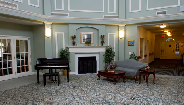 A spacious living room area with a patterned carpet, a black grand piano with a matching bench, a gray upholstered sofa, a wooden coffee table with a floral arrangement, and a fireplace with decorative plants and a framed picture above it. The walls are painted light green with white trim, and there are two wall sconces providing light. To the right, there is a hallway with carpeted flooring and ceiling lights.