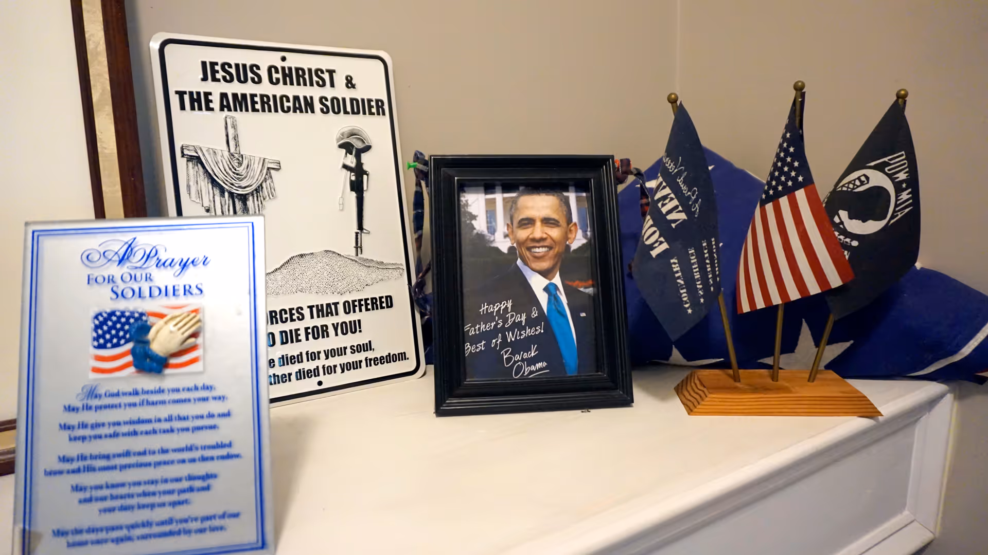 A display on a white surface featuring a framed photo of Barack Obama with a Father's Day message, a sign titled 'Jesus Christ & The American Soldier' with a cross and helmet illustration, a prayer card for soldiers with an American flag and praying hands, and a wooden stand holding three small flags including the American flag and POW-MIA flags.
