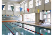 Indoor swimming pool with lane dividers, colorful triangular flags hanging above, large windows letting in natural light, and a small table with chairs near the poolside.